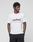 A man stands against a gray background, hands in pockets, wearing the LIVE FAST DIE YOUNG Basic Live Fast Graffiti Tee—a regular fit white T-shirt with a bold black "LiveFast" print and pink graffiti text. Made from 100% Baumwolle.
