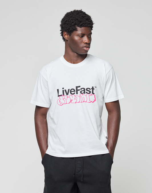 A man stands against a gray background, hands in pockets, wearing the LIVE FAST DIE YOUNG Basic Live Fast Graffiti Tee—a regular fit white T-shirt with a bold black "LiveFast" print and pink graffiti text. Made from 100% Baumwolle.