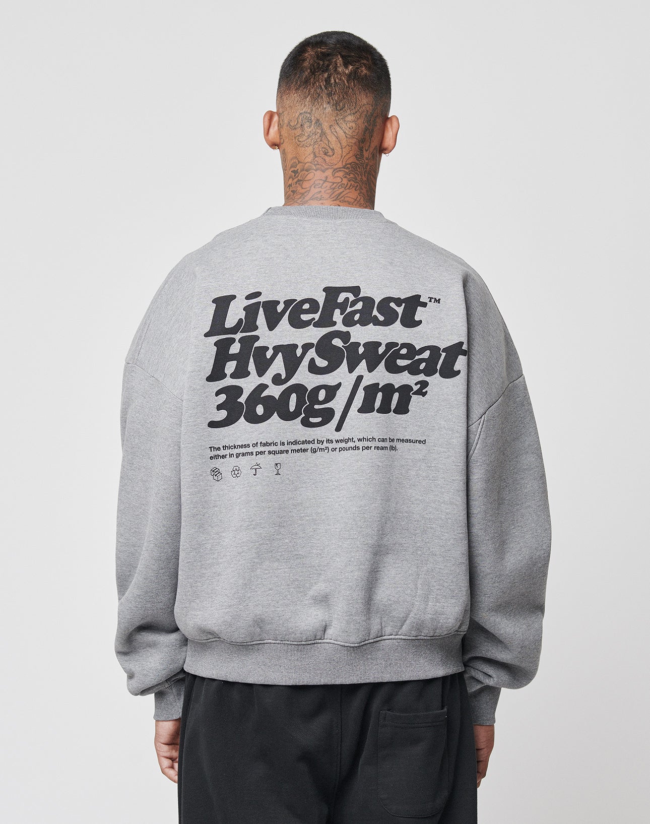 A person with short hair and neck tattoos wears the LIVE FAST DIE YOUNG Basic 360 Sweater, an oversize gray sweatshirt with bold black Live Fast Hvy Sweat 360g/m² backprint and subtle topstitching, set against a plain white background.
