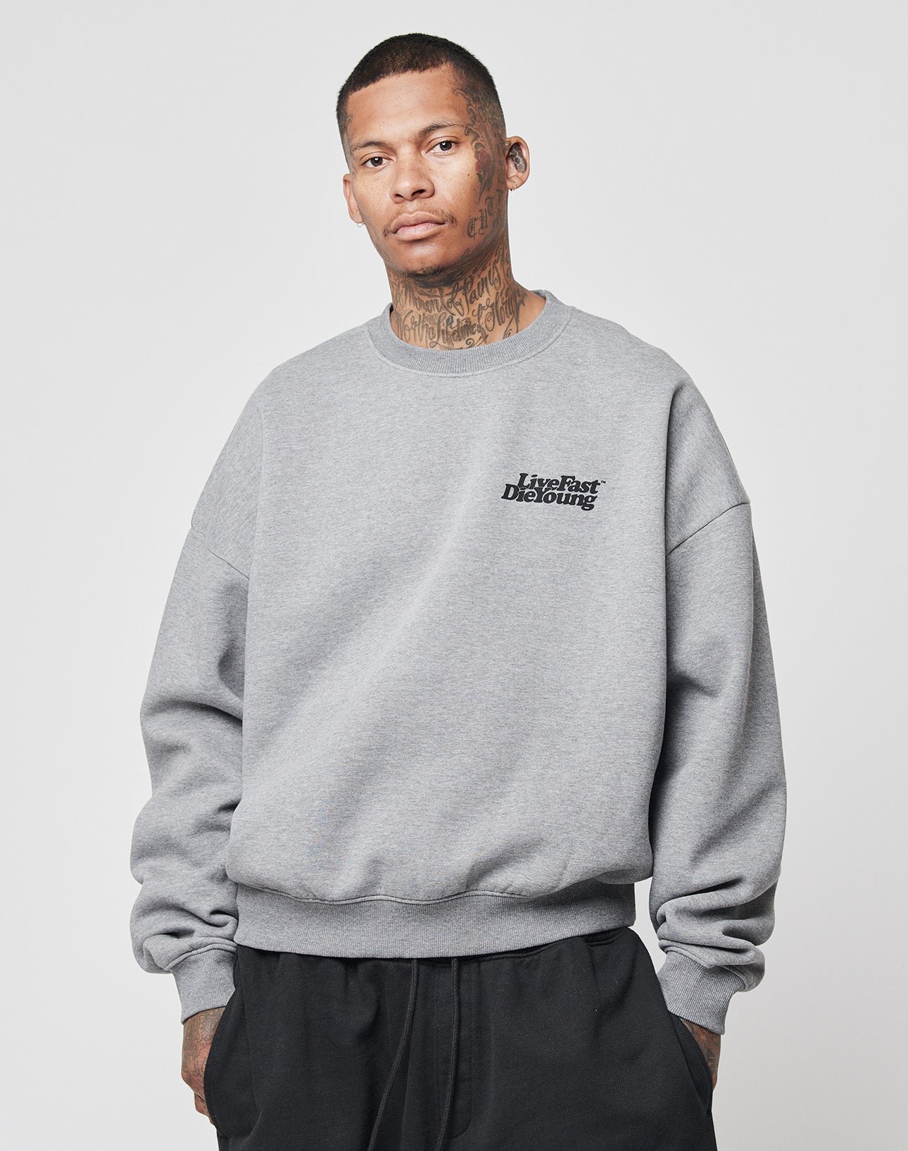 A person with tattoos wears the LIVE FAST DIE YOUNG Basic 360 Sweater—an oversize gray sweatshirt with front and back prints—paired with black pants, standing before a white background and looking neutrally at the camera.