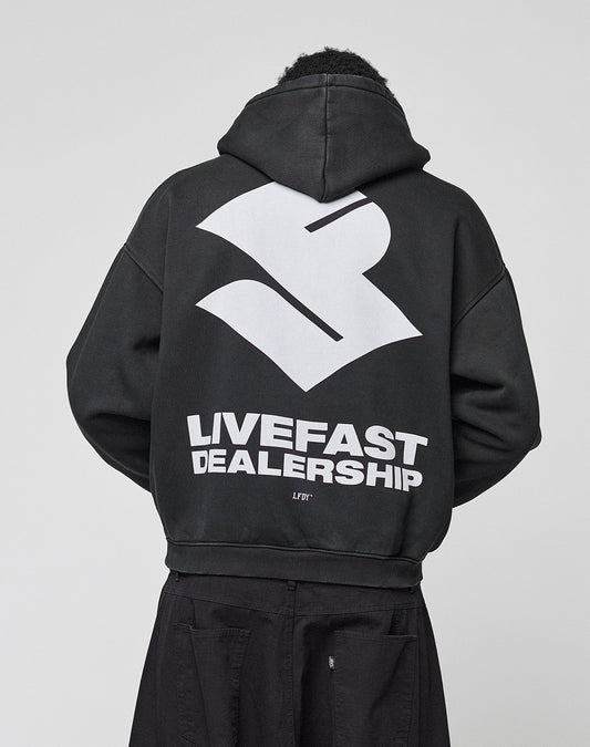 A person wears the LIVE FAST DIE YOUNG LFDY Dealership Hooded, an oversize black Baumwolle hoodie with a bold white abstract logo and "LIVEFAST DEALERSHIP" backprint, photographed from behind against a plain light background.