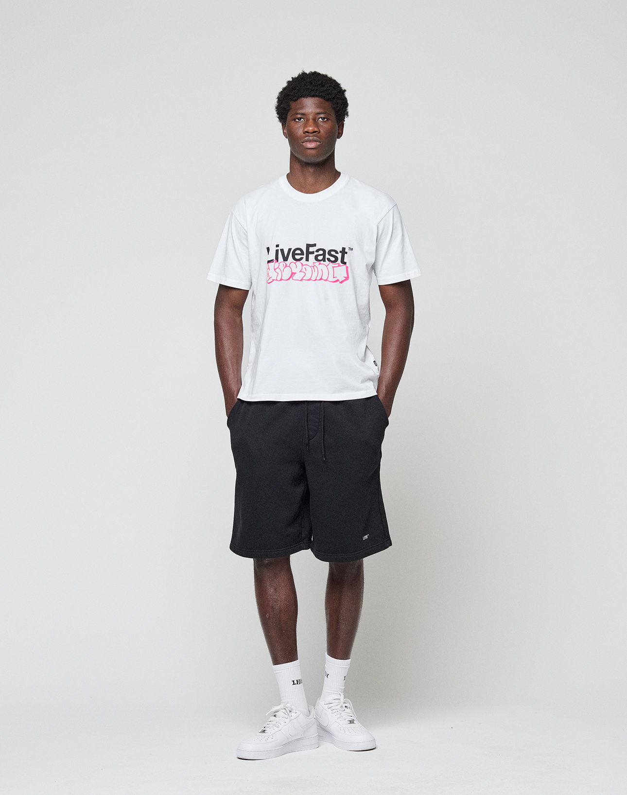 A man faces forward against a white background, wearing the LIVE FAST DIE YOUNG Basic Live Fast Graffiti Tee in regular fit (100% Baumwolle), black shorts, white socks, and white sneakers. His arms rest relaxed at his sides.