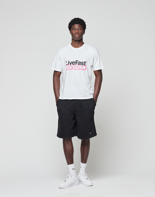 A man faces forward against a white background, wearing the LIVE FAST DIE YOUNG Basic Live Fast Graffiti Tee in regular fit (100% Baumwolle), black shorts, white socks, and white sneakers. His arms rest relaxed at his sides.