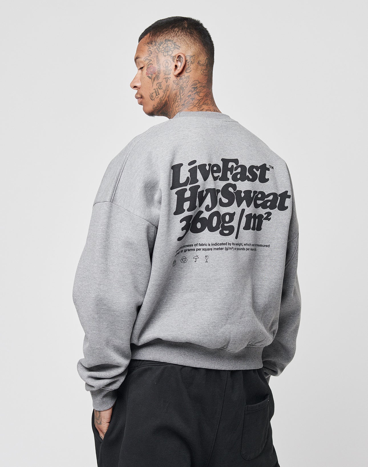 A tattooed person faces away wearing the LIVE FAST DIE YOUNG Basic 360 Sweater, an oversize gray sweatshirt with bold black “Live Fast Hvy Sweat 360g/m²” text on the back.