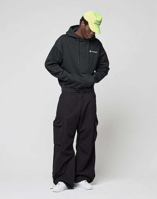 A person in a black LIVE FAST DIE YOUNG LFDY Dealership Hooded with front and back print, black cargo pants, white sneakers, and a neon green cap stands against a light background, hands in the hoodie pocket and gazing slightly down.