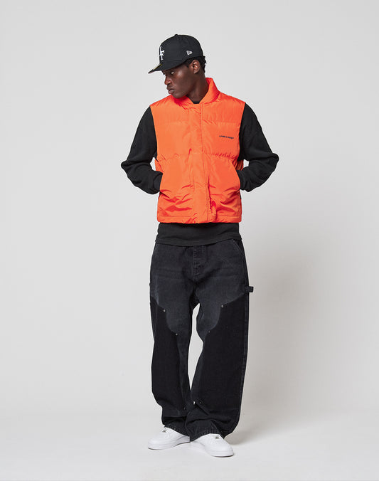 A person wears a black cap, black sweatshirt, white sneakers, black baggy jeans, and the bright orange "Live Fast Cropped Vest" by LIVE FAST DIE YOUNG, standing against a plain white background, looking to the side with hands in vest pockets.