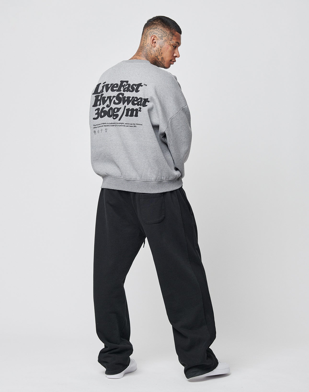 A man faces away from the camera, wearing the LIVE FAST DIE YOUNG Basic 360 Sweater—an oversized gray sweatshirt with black backprint text—and loose black pants, set against a plain white background.