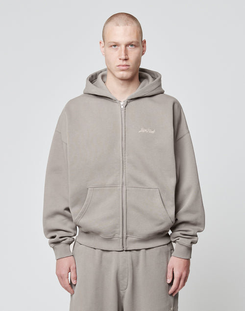 A person with a shaved head wears the LIVE FAST DIE YOUNG Basic Script Hooded Zip—a loose-fitting, gray Baumwolle-Polyester hoodie and sweatpants—standing against a light gray background, looking at the camera with a neutral expression.