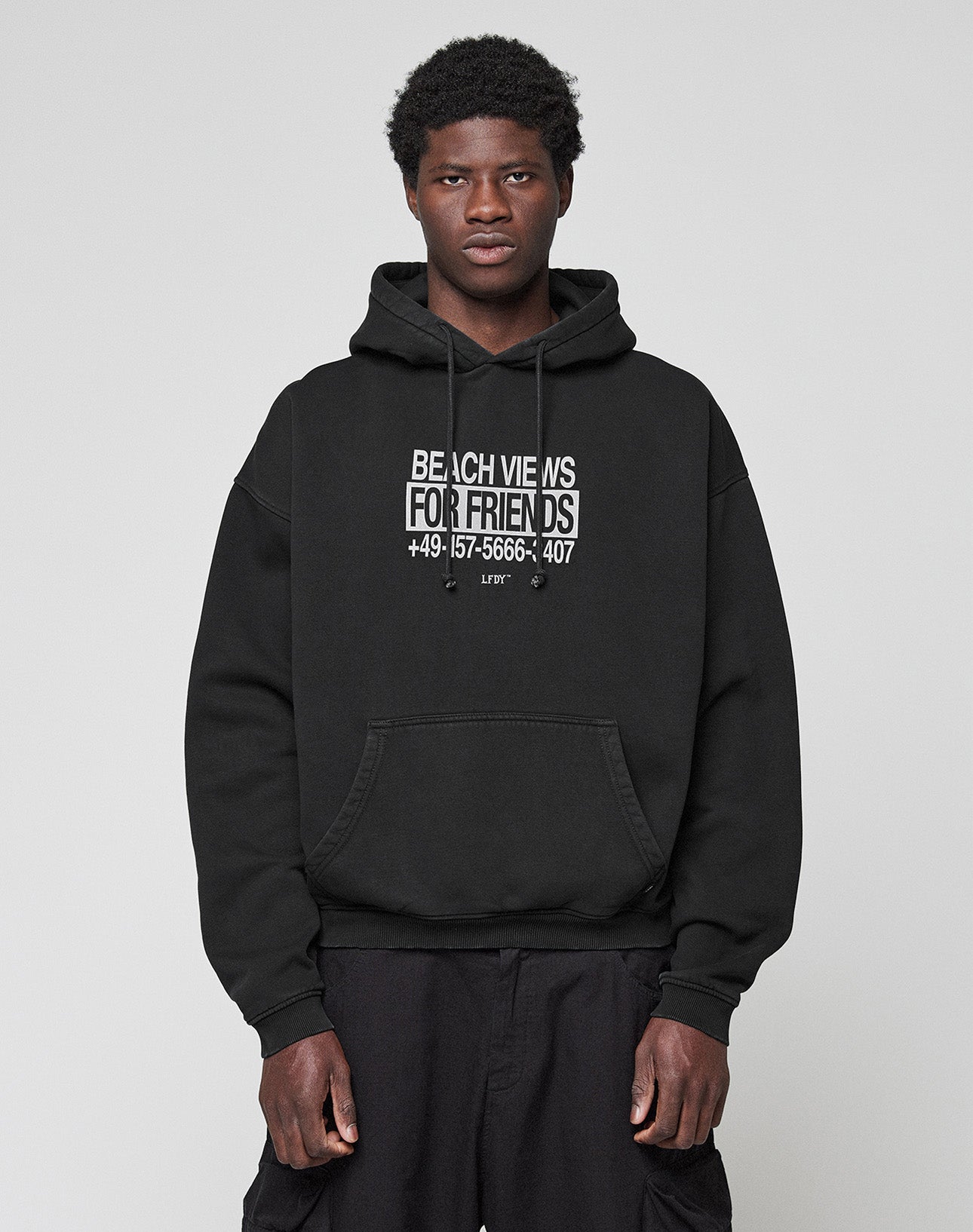 A person wears a black Live Fast Call Us Hooded by LIVE FAST DIE YOUNG, made of Baumwolle with BEACH VIEWS FOR FRIENDS +49-157-5666-3407 as bold front and back prints, standing against a plain light gray background.