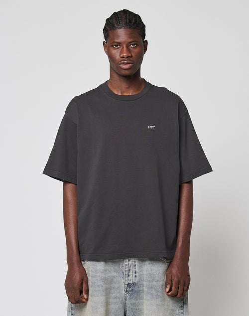 A man with braided hair models the LFDY Monaco Boys Tee—an oversize fit black T-shirt with small white chest text—paired with light blue jeans, standing against a plain light gray background.