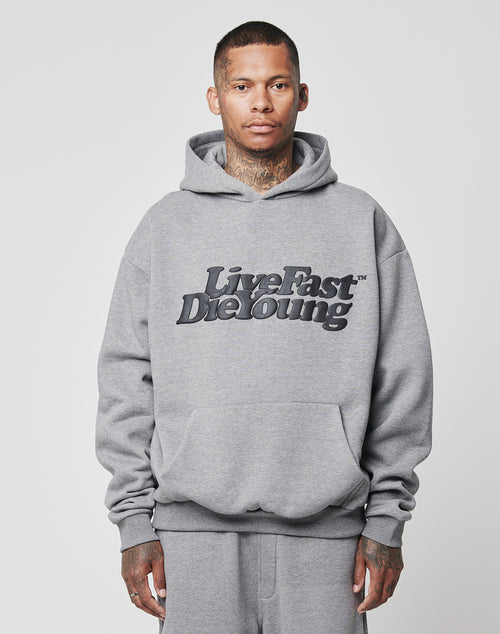 A man with short hair and visible neck tattoos wears a gray Basic Hooded sweatshirt by LIVE FAST DIE YOUNG, featuring the brand name in bold black letters. He stands against a plain, light gray background.