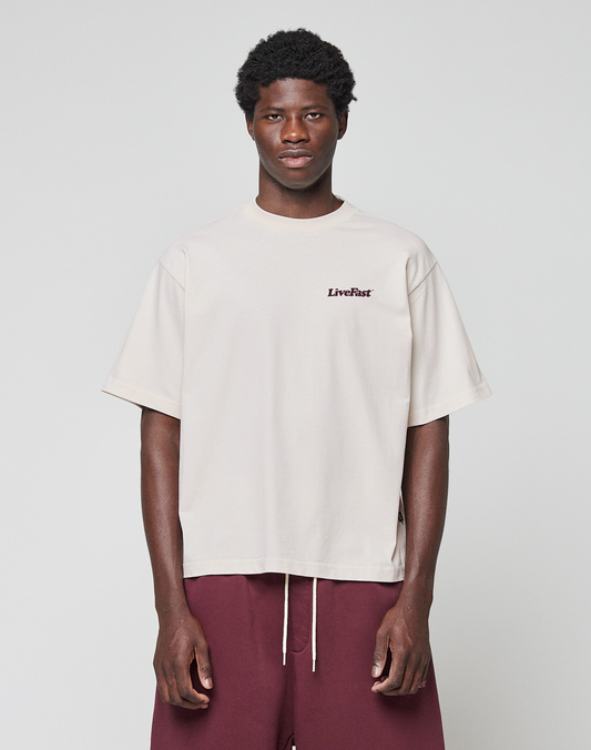 A person stands against a plain background wearing the LIVE FAST DIE YOUNG Basic 210 Tee in loose, light beige oversize fit with a LiveFast front print and maroon shorts. Made from 100% Baumwolle. Their arms rest at their sides as they look at the camera.