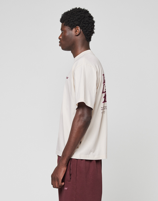 A person stands in profile facing left, wearing the LIVE FAST DIE YOUNG Basic 210 Tee in oversize cream with maroon front and back graphics and maroon shorts, set against a plain light gray background.