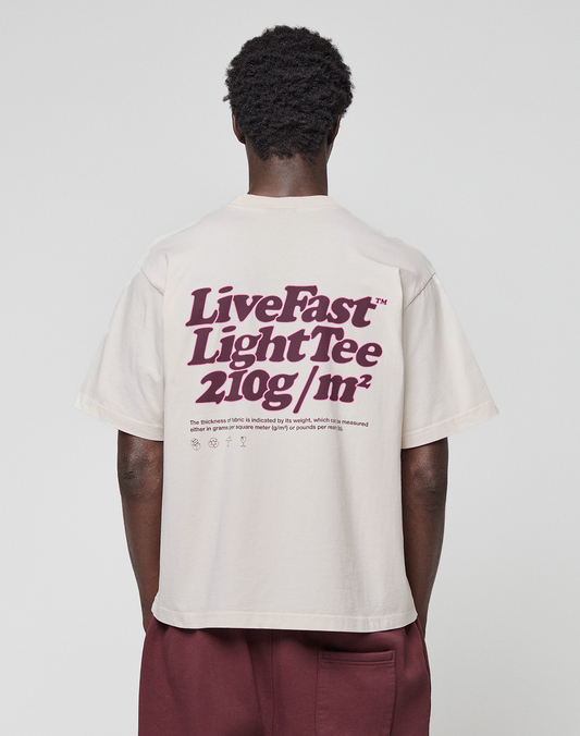 A person with short curly hair, seen from behind, wears a light beige LIVE FAST DIE YOUNG Basic 210 Tee featuring bold maroon text and small symbols, paired with maroon pants. The T-shirt is oversize fit and 100% Baumwolle.
