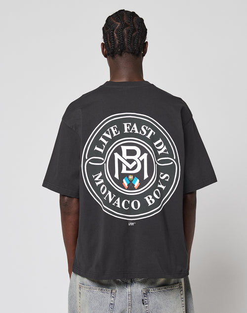 A person with braided hair, facing away, wears the LFDY Monaco Boys Tee—an oversized black T-shirt with a vintage effect and a large circular white LIVE FAST D1 MONACO BOYS logo plus colored emblem on the back.