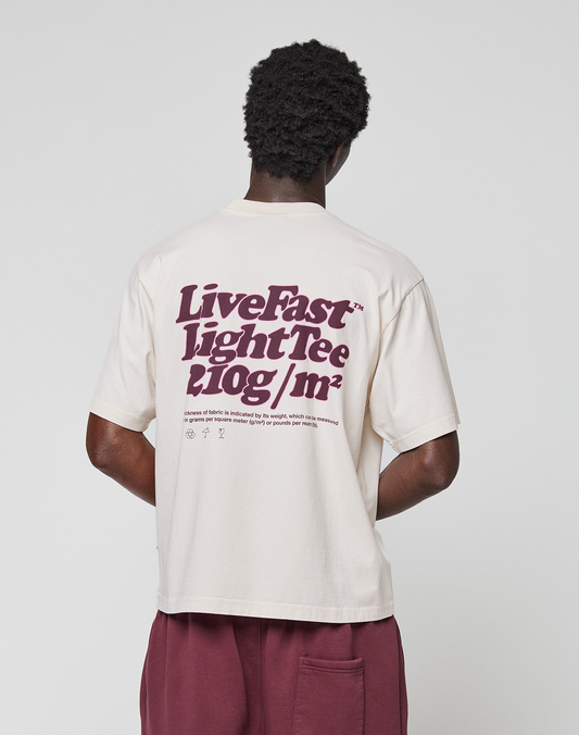 A person faces away, wearing an oversized cream Basic 210 Tee by LIVE FAST DIE YOUNG, made from 100% Baumwolle with a bold maroon back print and small text. The look is finished with maroon shorts.