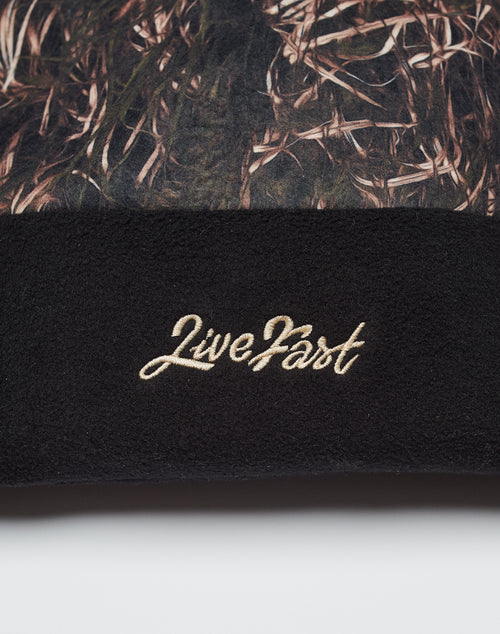 A close-up of the LIVE FAST DIE YOUNG Live Fast Gymbag reveals brown and green camo fabric on top, black fleece on the bottom, and LiveFast embroidered in beige script on the black polyester section.