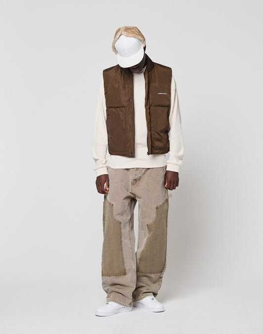 Wearing a white cap, cream sweatshirt, Live Fast Cropped Vest by LIVE FAST DIE YOUNG, loose tan patchwork jeans, and white sneakers, a person stands against a plain light background, looking down with hands by their sides.