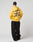 A person models the LIVE FAST DIE YOUNG Print Over Hooded in yellow, featuring bold black front and back prints, styled with black wide-leg pants, white shoes, and a black cap against a plain light backdrop.