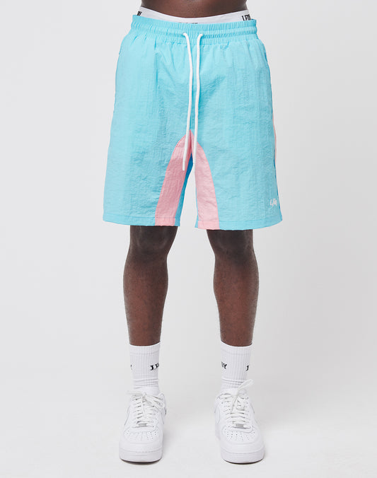 Only the lower half of a person is visible as they stand against a white background, wearing LIVE FAST DIE YOUNG Beach Shorts in light blue with pink accents and mesh inlay, paired with white crew socks and white sneakers.
