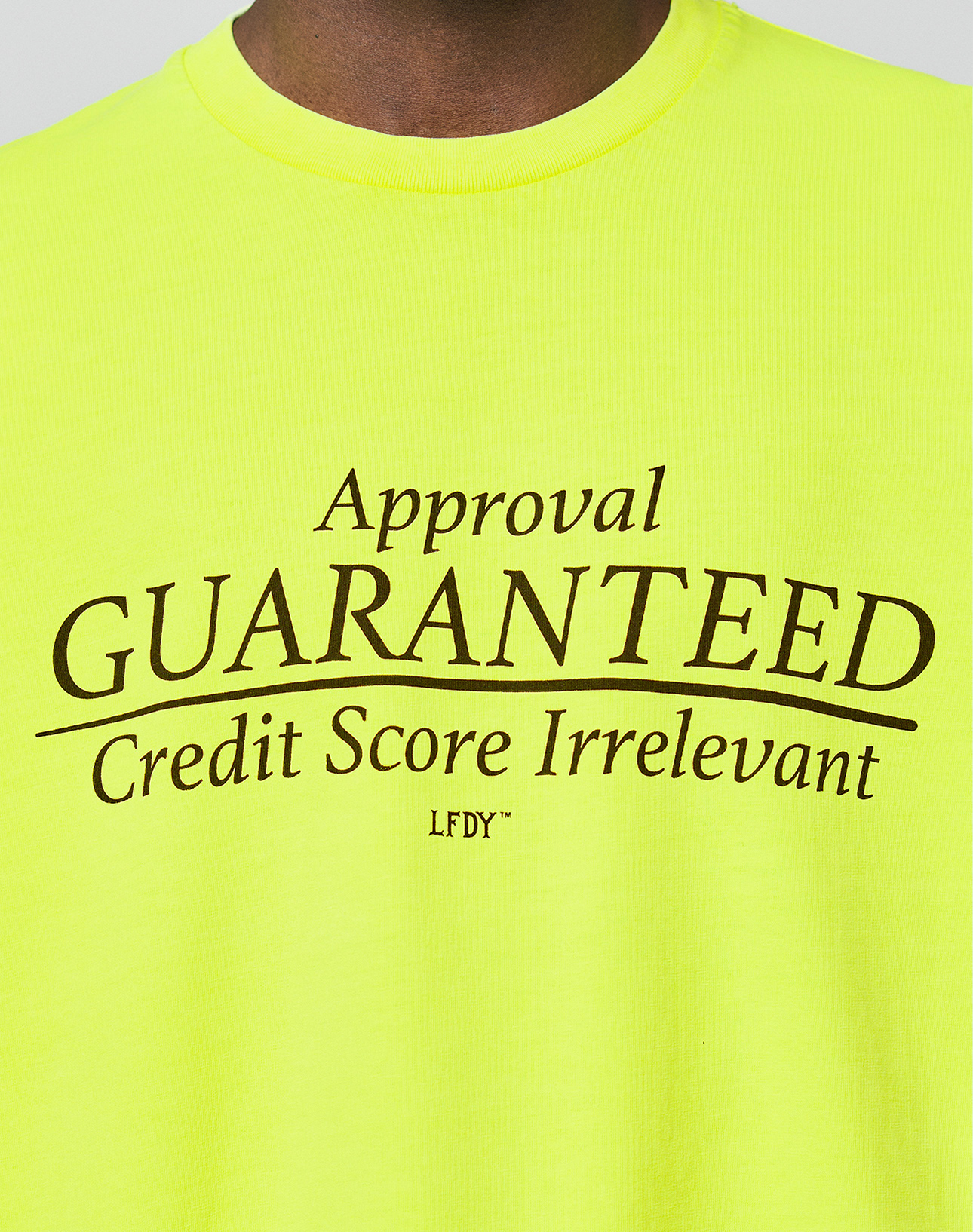 A person wears the LFDY Real Est.8 Longsleeve by LIVE FAST DIE YOUNG, an oversized bright yellow shirt with "Approval GUARANTEED Credit Score Irrelevant LFDY" boldly printed in black on the front, made from 100% Baumwolle for comfort.