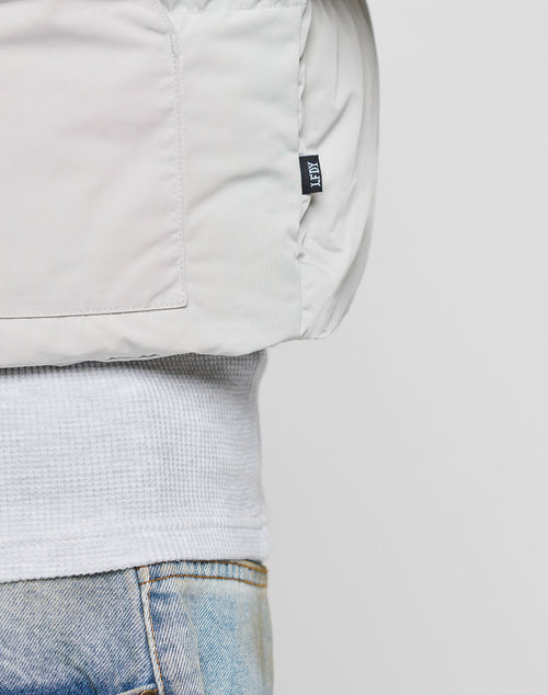 Close-up of a person wearing the LIVE FAST DIE YOUNG Basic Cropped Vest in light grey over a white textured shirt and light blue jeans. A small black brand tag appears on the vest’s side seam.