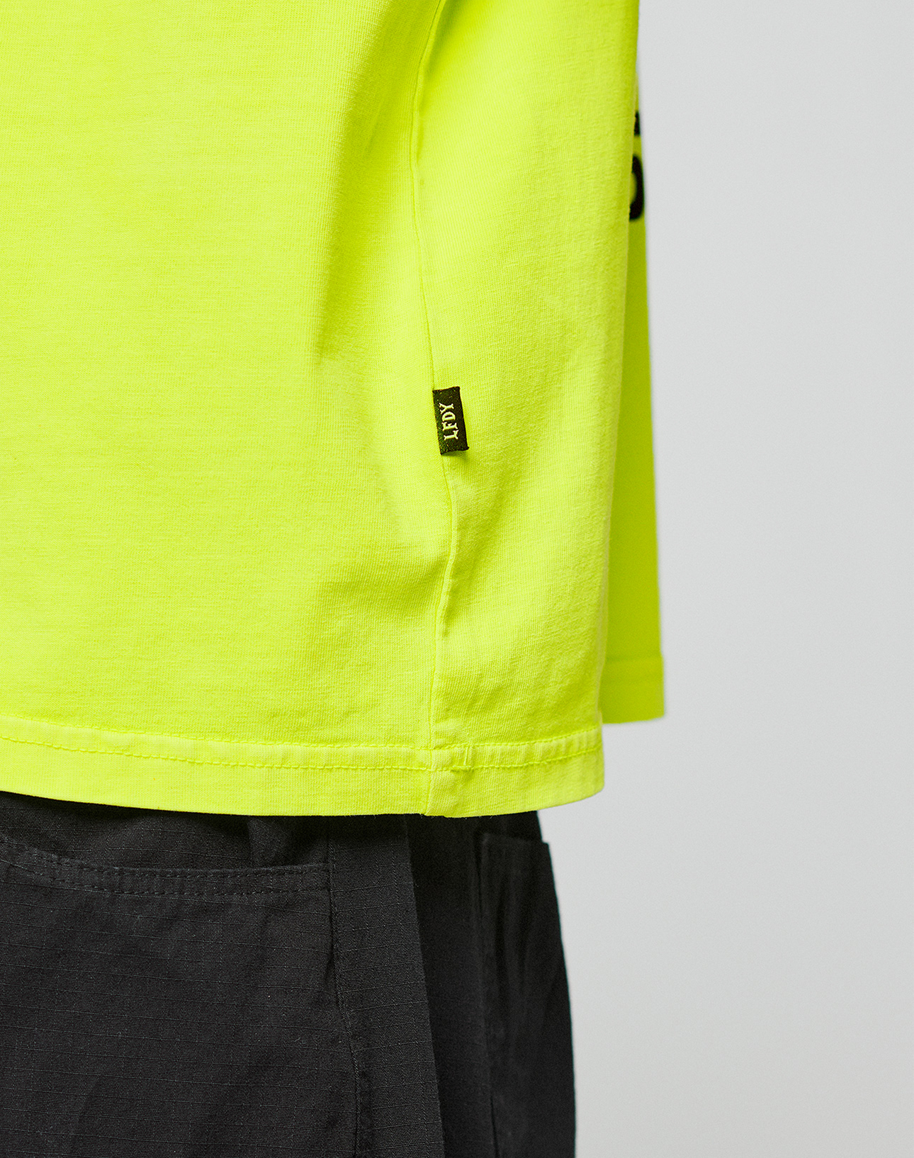 A close-up of a person wearing the LIVE FAST DIE YOUNG LFDY Real Est.8 Longsleeve in bright yellow with oversize fit and small black side tag, paired with dark gray pants, showing only the lower shirt and upper pants.
