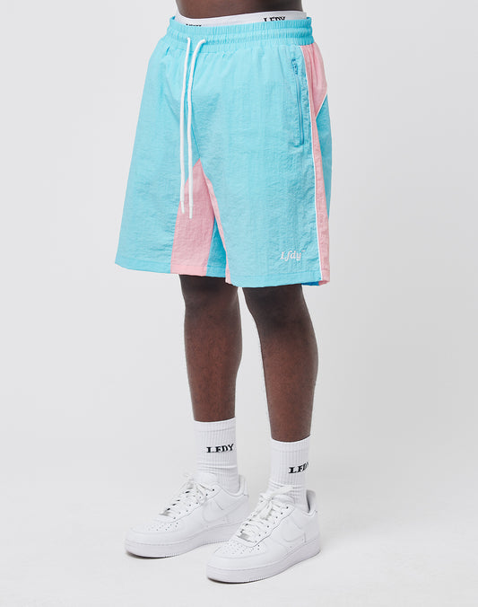 A person in LIVE FAST DIE YOUNG Beach Shorts in pastel blue and pink colorblock with mesh inlay, white socks with black LDXY text, and white sneakers stands against a plain white background.