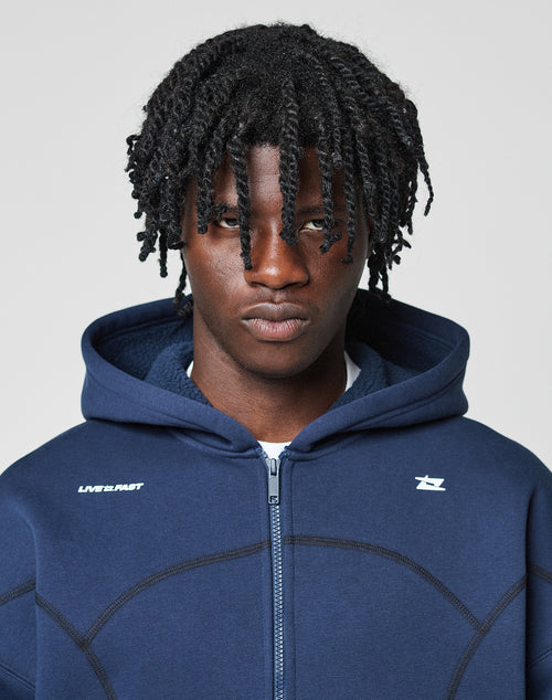 A person with short, twisted hair wears the LFDY Active Fleece Hooded Zip by LIVE FAST DIE YOUNG in navy blue, featuring an oversize fit, high collar, and white logo print, looking serious against a plain light background.
