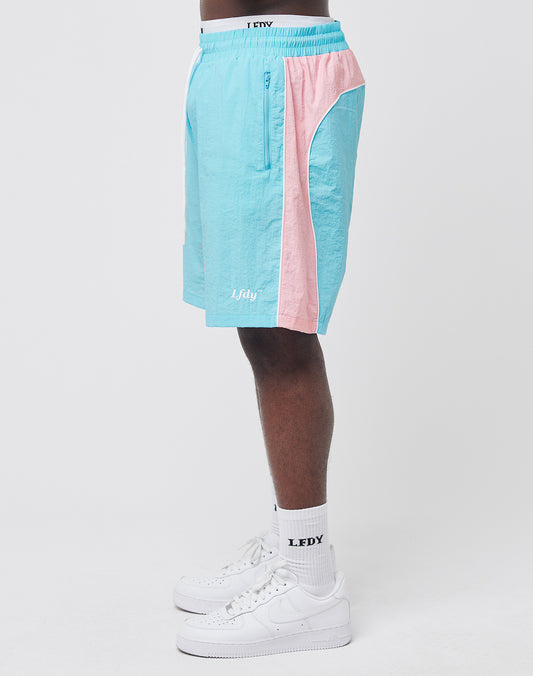 A person in profile wears LIVE FAST DIE YOUNG Beach Shorts in light blue and pink with mesh inlay, July printed on the leg, zippered side pockets, plus white LFDY socks and white Nike sneakers against a plain light background.