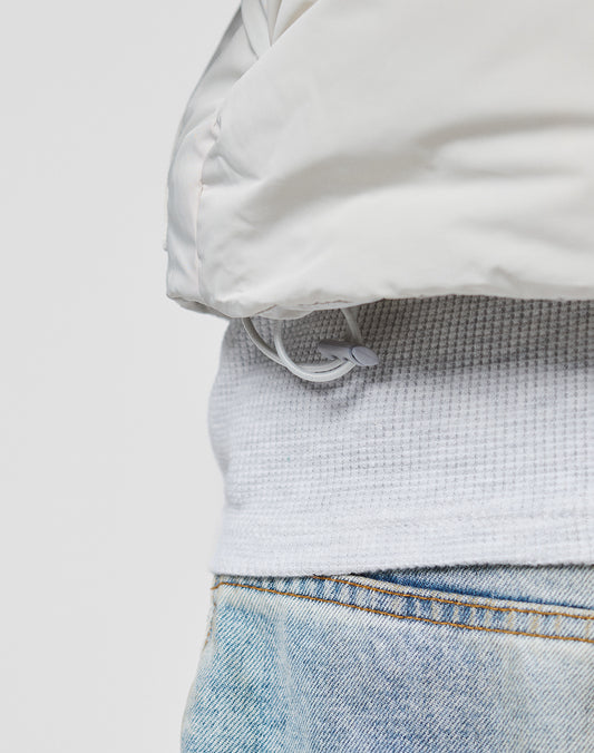 Close-up of a person wearing the LIVE FAST DIE YOUNG Basic Cropped Vest, light blue denim jeans, and a regular fit white puffer jacket with a drawstring hem. The background is plain and light-colored.