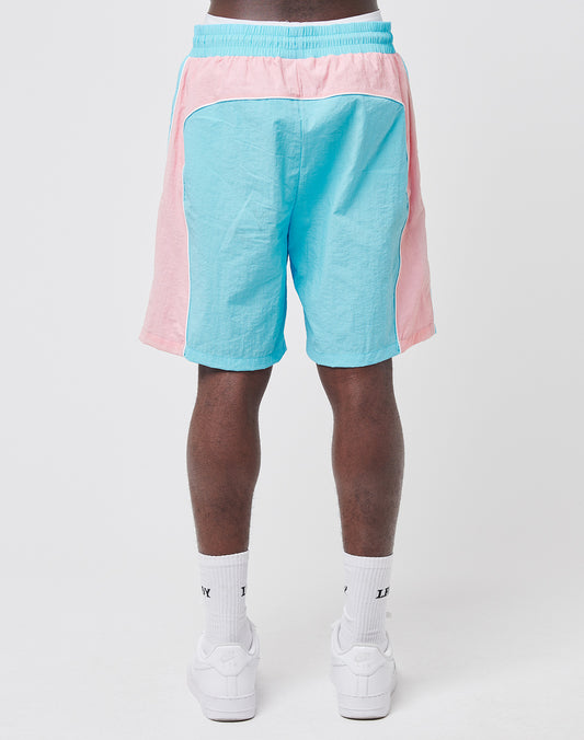A person faces away, wearing LIVE FAST DIE YOUNG Beach Shorts in light blue and pink colorblock with mesh inlay details, paired with white crew socks and sneakers against a plain white background.