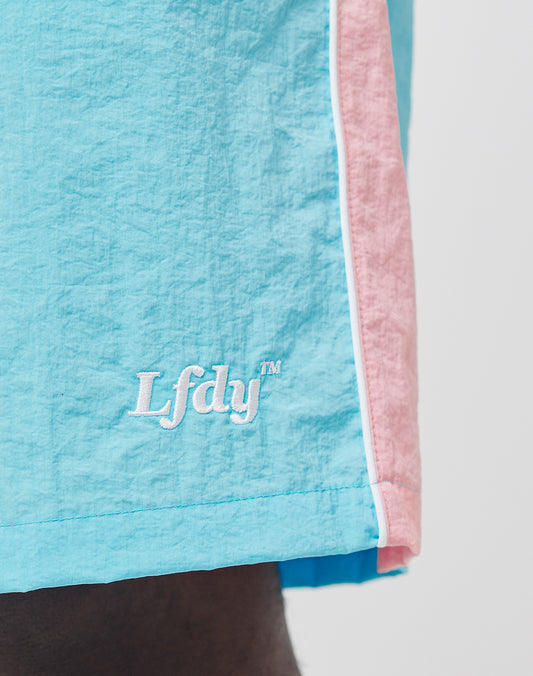 Close-up of LIVE FAST DIE YOUNG Beach Shorts in light blue and pink, featuring white piping, a mesh inlay, and “LFDY™” embroidered in white on the blue section.