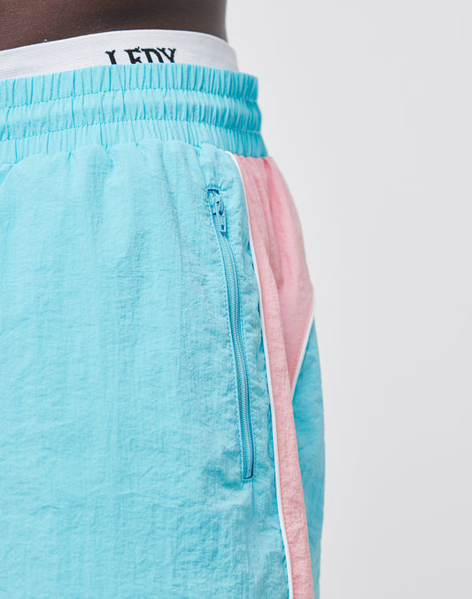 Close-up of a person wearing LIVE FAST DIE YOUNG Beach Shorts in light blue and pink color-block, featuring a regular fit, visible zipper pocket, elastic waistband, and mesh inlay. Part of their dark-skinned waist and torso is shown against a plain background.