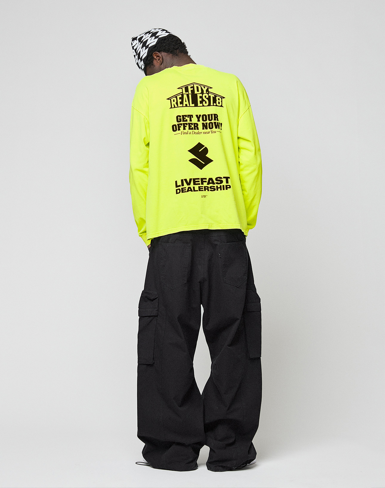 A person stands with their back to the camera, wearing a neon yellow LFDY Real Est.8 Longsleeve by LIVE FAST DIE YOUNG, featuring bold black front and back prints, loose black cargo pants, and a black-and-white checkered hat.