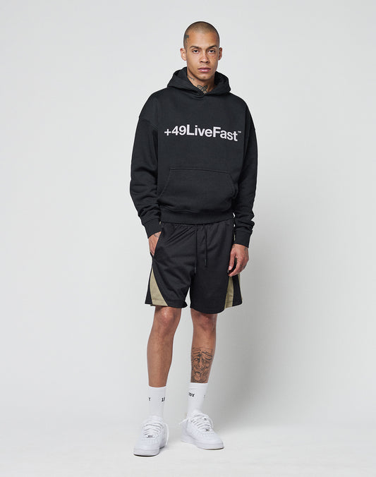 A man wears the LIVE FAST DIE YOUNG Basic +49 Hooded in black, featuring a +49LiveFast front print and oversize fit (Baumwolle-Polyester blend), paired with black shorts with beige side panels, white socks, and white sneakers.