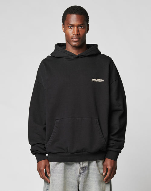 A person stands facing forward wearing an oversize fit black LFDY Savior Hooded sweatshirt by LIVE FAST DIE YOUNG, featuring a small white logo on the upper left chest, made from 100% Baumwolle, paired with light blue jeans. The background is plain and light.