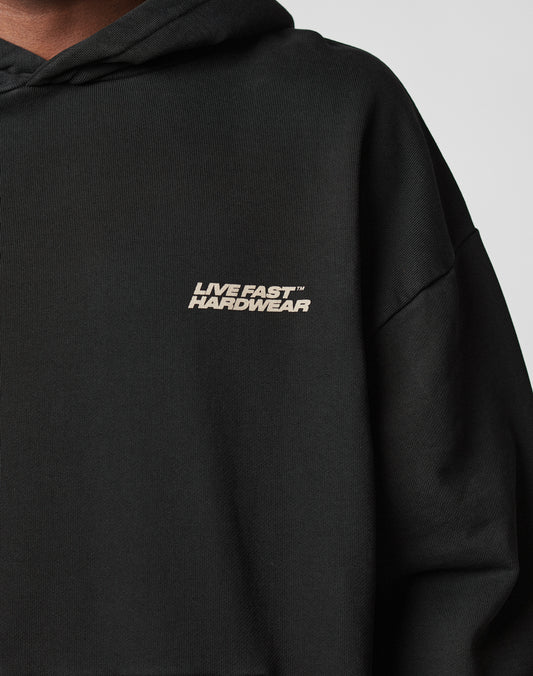 Close-up of a person wearing the LIVE FAST DIE YOUNG LFDY Savior Hooded in black, featuring an oversize fit and white LIVE FAST™ HARDWEAR print on the left chest. Made from 100% Baumwolle, hood up, upper body partially visible.