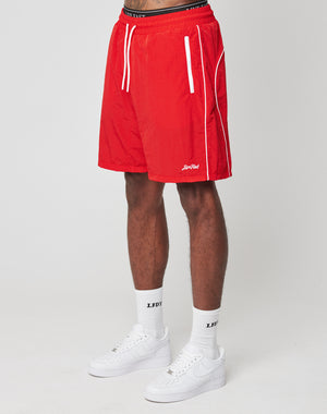 A person wears LIVE FAST DIE YOUNG Beach Shorts in bright red with white piping and an elastic waistband, paired with white crew socks featuring black letters and white Nike sneakers, standing against a plain light background. Only the lower body is shown.