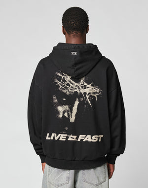 A person in the LFDY Savior Hooded by LIVE FAST DIE YOUNG, an oversize black 100% Baumwolle hoodie with a bold face graphic, abstract designs, and LIVE FAST on the back, stands against a plain light background.