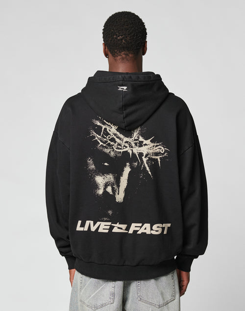 A person in the LFDY Savior Hooded by LIVE FAST DIE YOUNG, an oversize black 100% Baumwolle hoodie with a bold face graphic, abstract designs, and LIVE FAST on the back, stands against a plain light background.