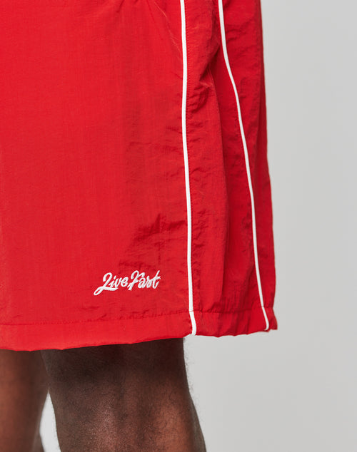 Close-up of a person wearing bright red LIVE FAST DIE YOUNG Beach Shorts with an Elastik-Bund and white piping. Live Fast is embroidered near the hem. The background is plain and light-colored.