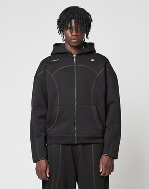 A person with medium-length twisted hair wears the LIVE FAST DIE YOUNG LFDY Active Fleece Hooded Zip in black, styled oversized with matching black pants, standing against a plain light gray background and looking at the camera.