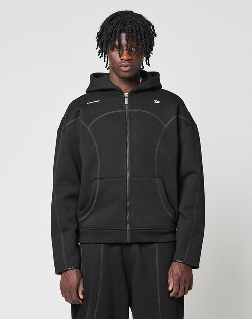 A person with medium-length twisted hair wears the LIVE FAST DIE YOUNG LFDY Active Fleece Hooded Zip in black, styled oversized with matching black pants, standing against a plain light gray background and looking at the camera.