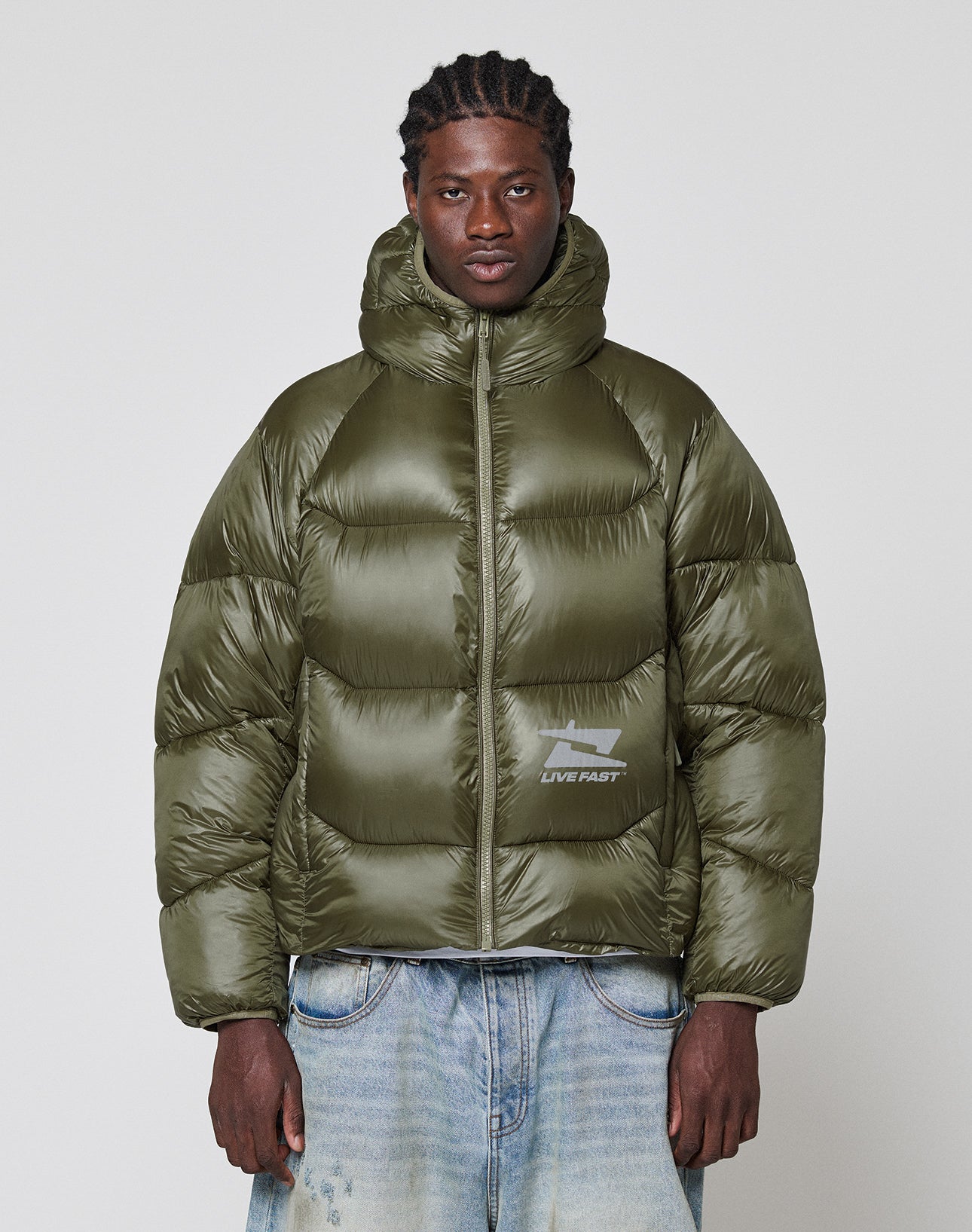 A man wears the LIVE FAST DIE YOUNG Live Fast Puffer Jacket in olive green with nylon outer material, oversized fit, hood, and a reflective LIVE FAST logo, styled with light blue jeans against a plain background.