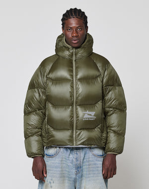 A man wears the LIVE FAST DIE YOUNG Live Fast Puffer Jacket in olive green with nylon outer material, oversized fit, hood, and a reflective LIVE FAST logo, styled with light blue jeans against a plain background.