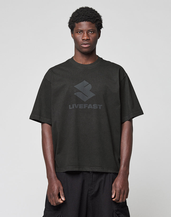 A person stands against a light gray background, wearing the Live Fast LF Tee by LIVE FAST DIE YOUNG—an oversize-fit black T-shirt with a "LIVEFAST" front print, made from 100% Baumwolle—and paired with black shorts.