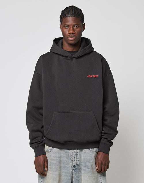 A person faces forward in a LIVE FAST DIE YOUNG LFDY Globe Hooded black hoodie with red LIVE FAST text on the left chest, made of cotton-polyester, and light blue jeans. The background is plain light gray.