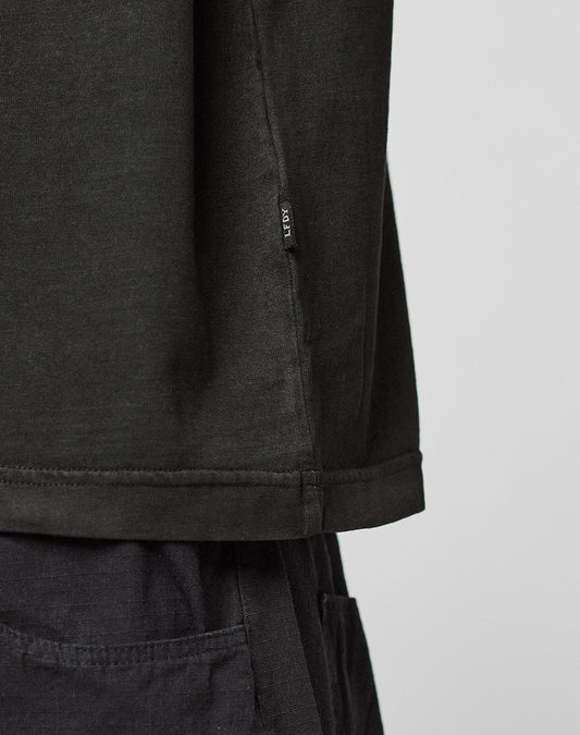 Close-up of a person wearing the Live Fast LF Tee by LIVE FAST DIE YOUNG, an oversized black shirt with black pants. A small fabric tag is attached near the hem. The background is plain and light-colored.