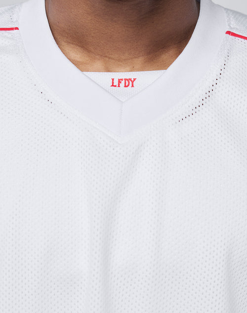 Close-up of a person wearing the Basic Mesh Jersey by LIVE FAST DIE YOUNG. This white oversize-fit sports jersey is made from 100% polyester with a V-neck collar. Red LFDY letters are visible inside the collar; face not shown.